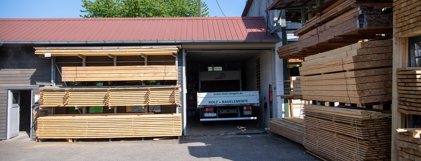 Gestapelte Bauholzlatten links und rechts vor Lagerhalle bei Holz Langen.