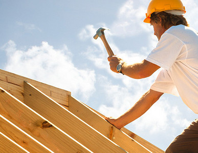 Dachdecker mit gelbem Schutzhelm beim Befestigen von Konstruktionsvollholz (KVH) auf einem Dachstuhl unter strahlendem Himmel.