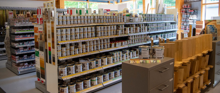 SB-Bereich bei Holz Langen mit Regalen voller Holzfarben und -öle sowie ausgestellten Holzmusterplatten.