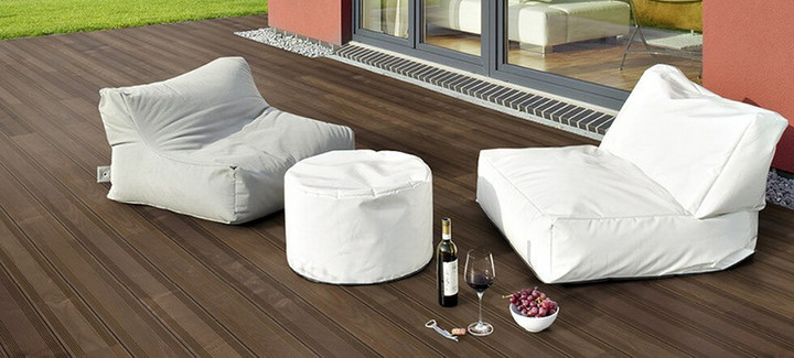 Dunkle Holzterrasse neben einem Gebäude mit zwei Sitzkissen und dekorativen Elementen.