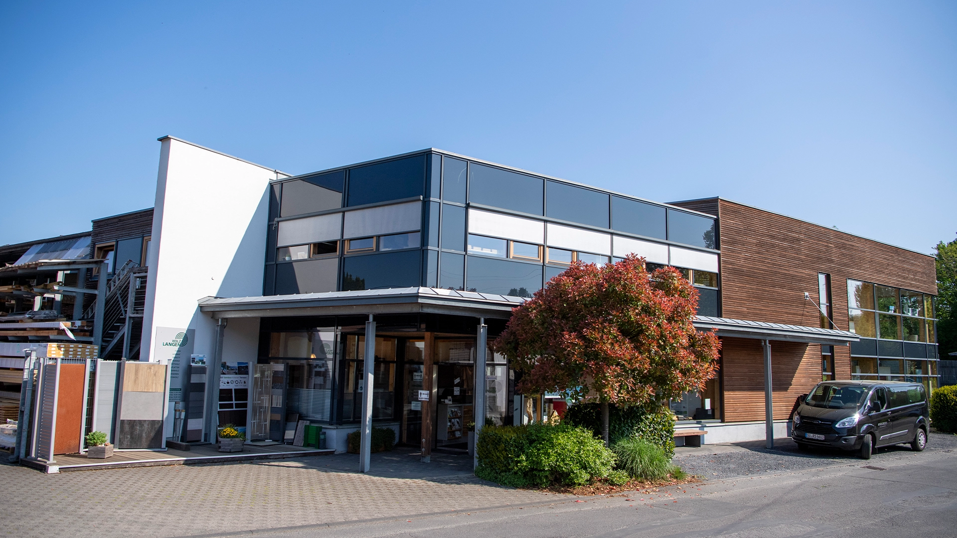 Außenansicht des Holz-Langen-Gebäudes mit großer Glasfassade, Holzelementen und Parkplatz im Vordergrund.