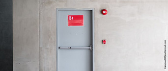 Graue Brandschutztür mit Fluchtwegsymbol und roter Alarmglocke.